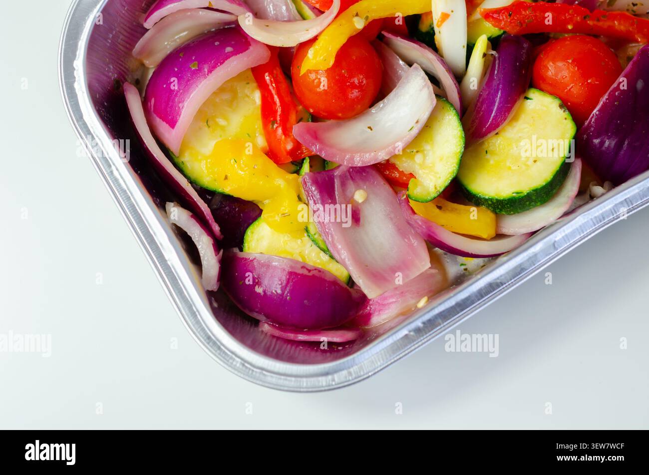 Les légumes vibrants dans un plateau métallique brillent sous une lumière vive. Les oignons rouges, les courgettes, les poivrons et les tomates cerises remplissent le cadre. Frais, coloré, an Banque D'Images