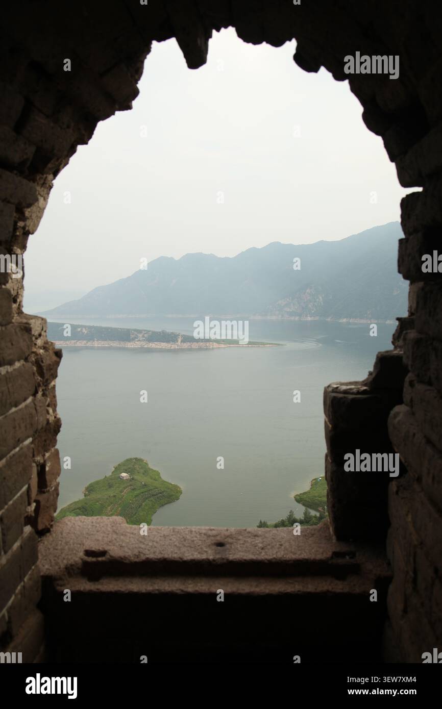 Encadré par l'arc brisé d'une tour de guet de la Grande Muraille, un paysage serein se déploie : un lac calme s'étend sous un ciel brumeux, sa surface bleu ardoise b Banque D'Images