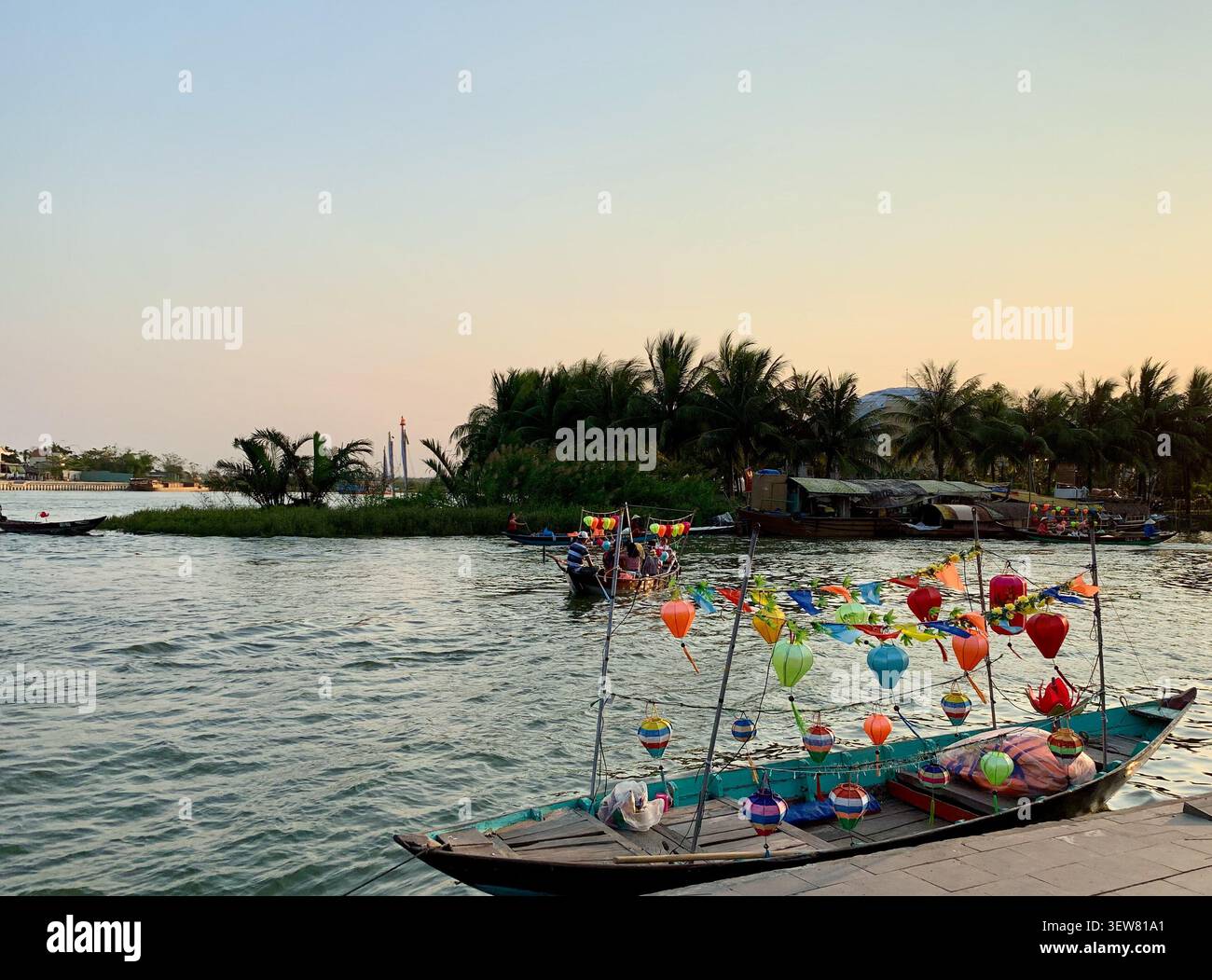 Bateau traditionnel coloré en bois décoré de lanternes en soie amarré sur la rivière Thu bon au crépuscule dans la vieille ville de Hoi an, Vietnam, avec des palmiers - Image de stock capturée avec un smartphone