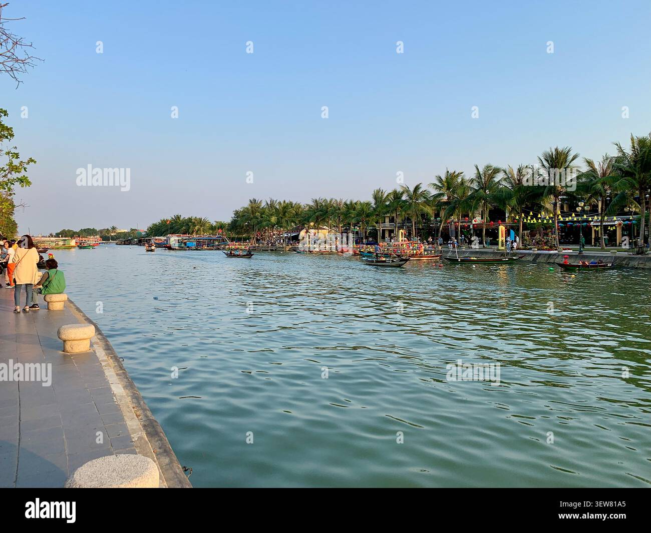 La promenade de la rivière Thu bon dans la ville antique de Hoi an, Vietnam, avec des bateaux traditionnels en bois amarrés le long du front de mer, des palmiers bordant la rive éloignée - Image de stock capturée avec un smartphone