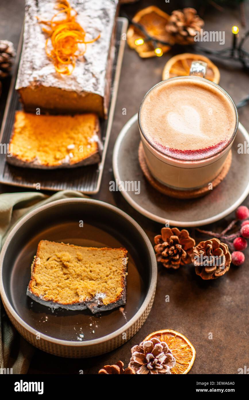 Un cadre de table avec un cappuccino chaud au cœur latte art, un gâteau des fêtes saupoudré entier et une tranche unique servie dans un bol en céramique sombre. Banque D'Images