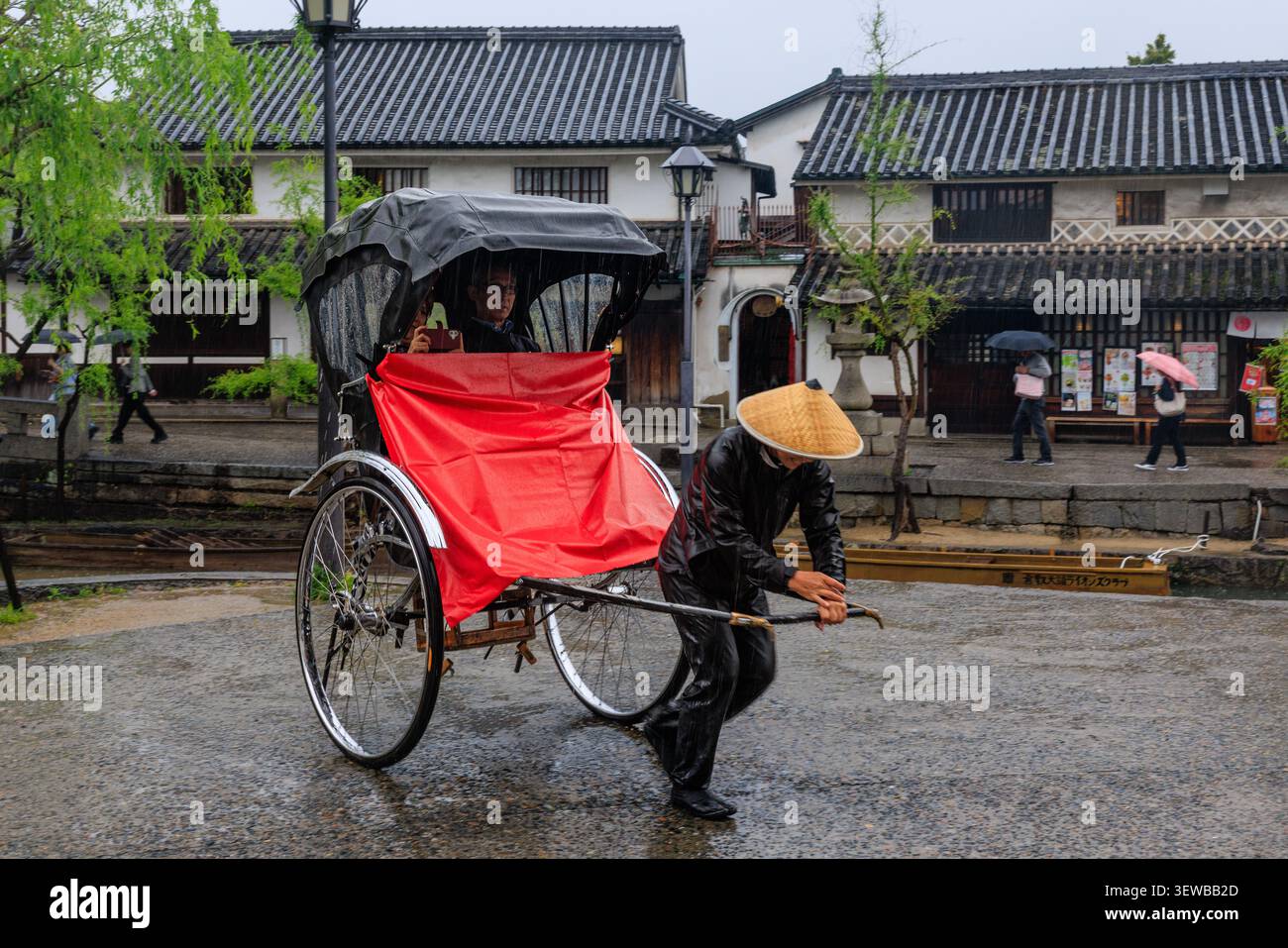 Une rickshaw japonaise traditionnelle avec un toit rouge vif est tirée par un conducteur à chapeau de paille le long d'une rue historique pluvieuse à Kurashiki Banque D'Images