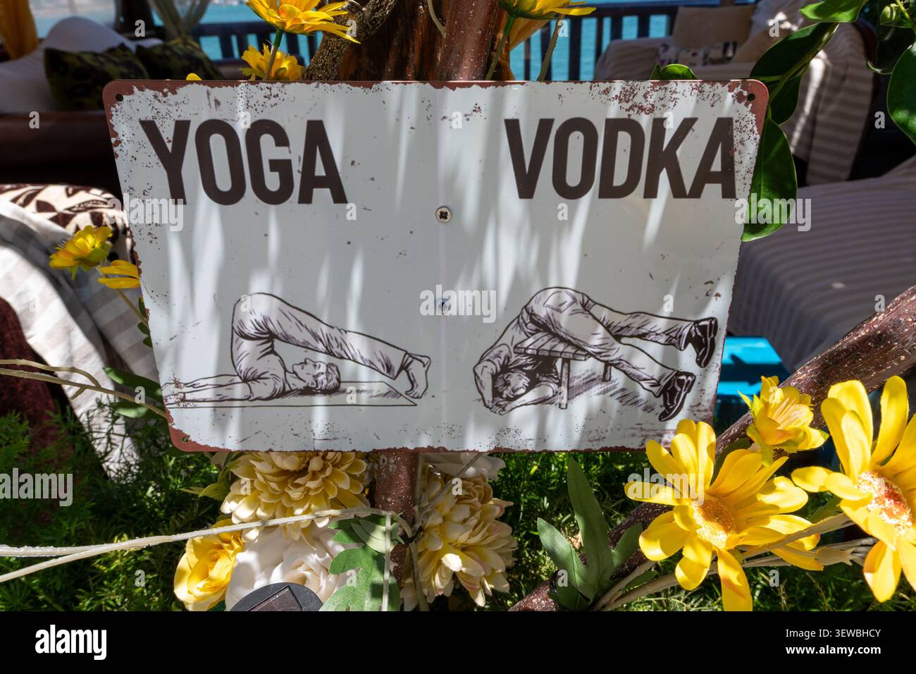 Un signe humoristique en métal blanc altéré avec un texte noir lisant Yoga et Vodka, illustrant un étirement de yoga à côté d'un homme ivre évanoui sous un bois Banque D'Images