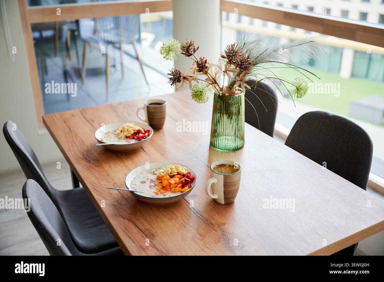 Petit déjeuner servi sur la table à manger, bols de céréales avec des fruits et des tasses de thé ou de café dans l'intérieur moderne de la maison, table en bois par fenêtre avec lumière naturelle, concept de routine matinale saine Banque D'Images