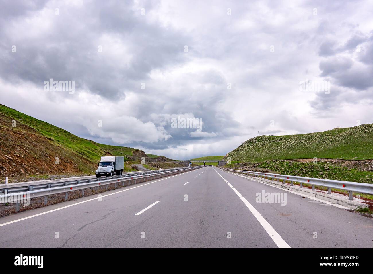 Large autoroute s'étend à travers des collines verdoyantes sous le ciel nuageux. Un seul camion blanc avance Banque D'Images
