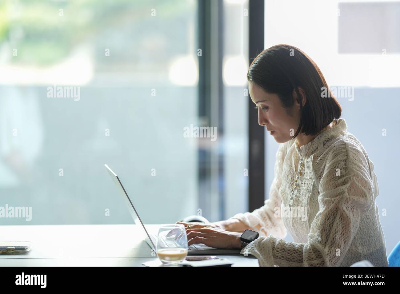 Une lumière douce brille à travers les grandes fenêtres. Femme s'assoit tranquillement, concentré sur l'écran de l'ordinateur portable. Ses doigts se déplacent doucement sur les touches du clavier. Le verre à café reste n Banque D'Images