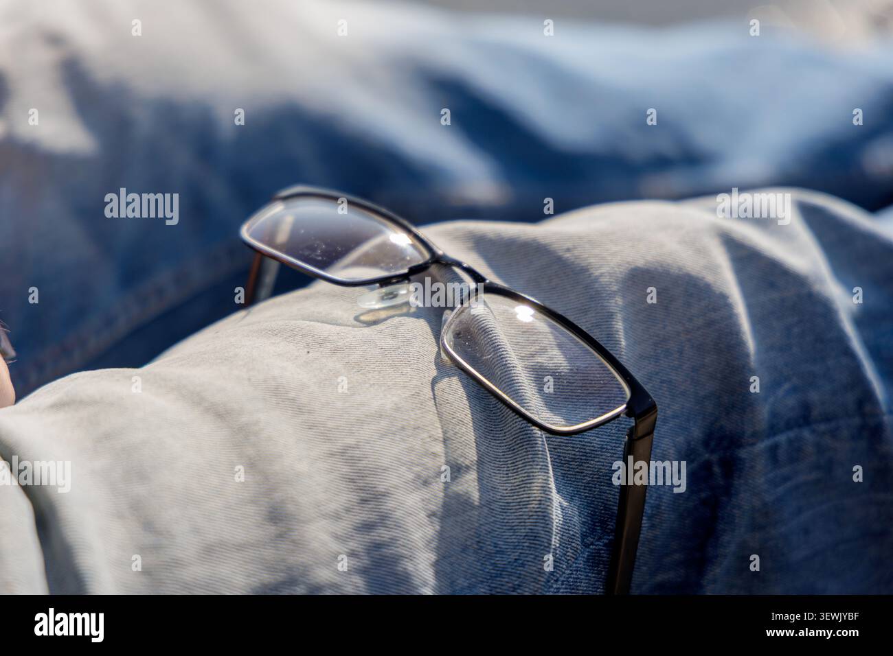 Gros plan de lunettes noires classiques reposant sur les genoux d'une personne portant un Jean denim bleu en plein air en plein soleil. Banque D'Images
