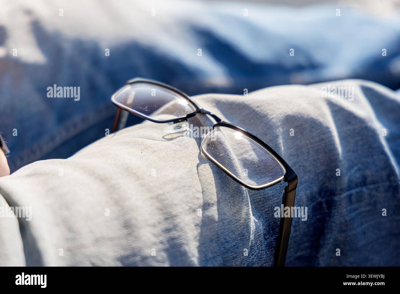 Gros plan de lunettes noires classiques reposant sur les genoux d'une personne portant un Jean denim bleu en plein air en plein soleil. Banque D'Images