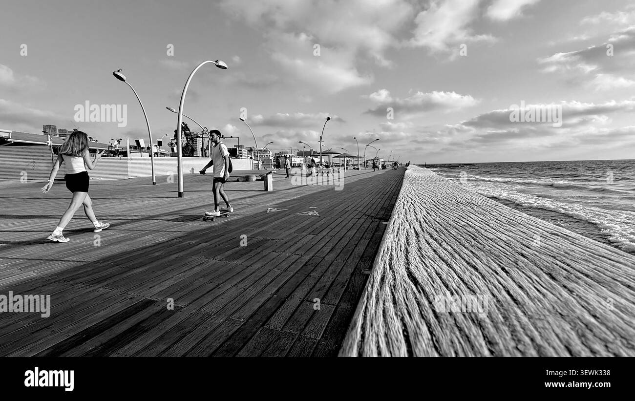 Une photographie grand angle en noir et blanc d'une promenade côtière. Les gens marchent et font du skateboarding le long de la passerelle en bois à côté de la mer, featu - Image de stock capturée avec un smartphone