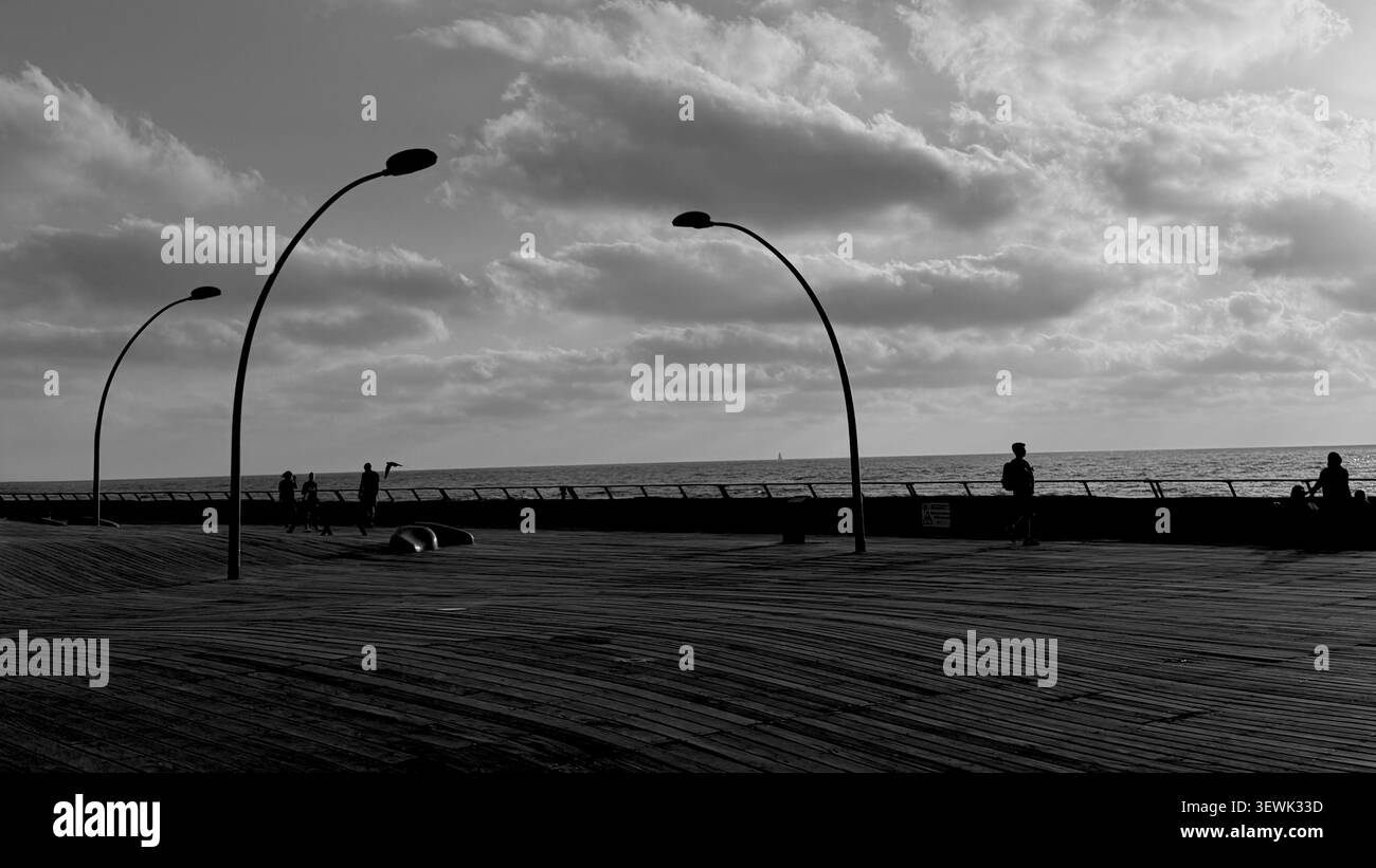 Une photographie en noir et blanc à grand angle capturant des poteaux de lampadaires incurvés et des silhouettes de personnes marchant le long d'une large promenade en bois côtière Banque D'Images