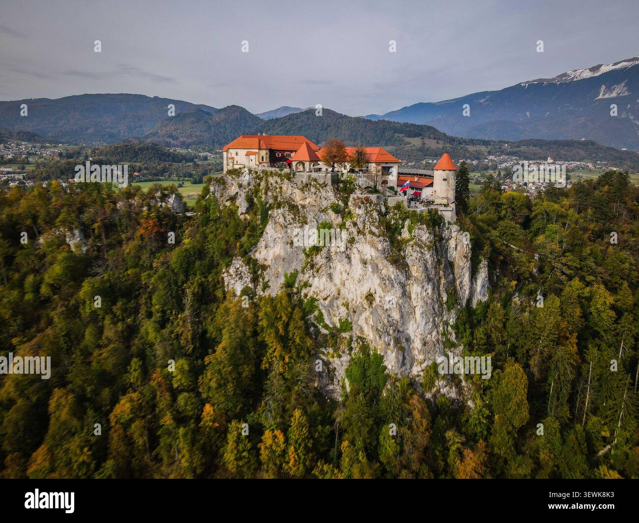 Bled Castle avec ses toits rouges se dresse au sommet d'une falaise rocheuse entourée d'arbres verts et oranges, avec des montagnes lointaines et une ville dans l'arrière-pays Banque D'Images