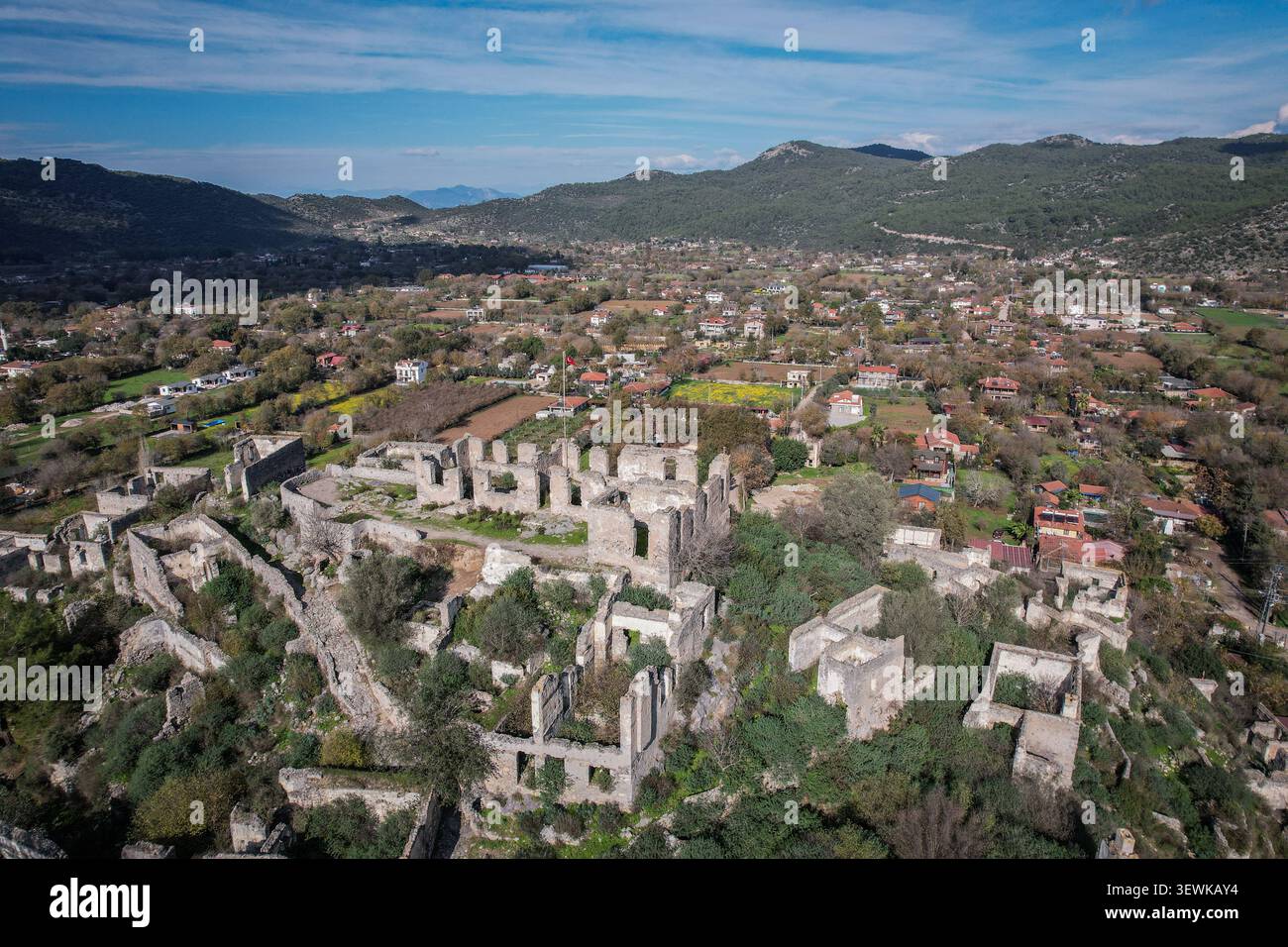 Une vue aérienne grand angle capture les ruines de pierre antiques de Kayakoy Ghost Town entouré d'une végétation verte, avec le village moderne et lointain h Banque D'Images