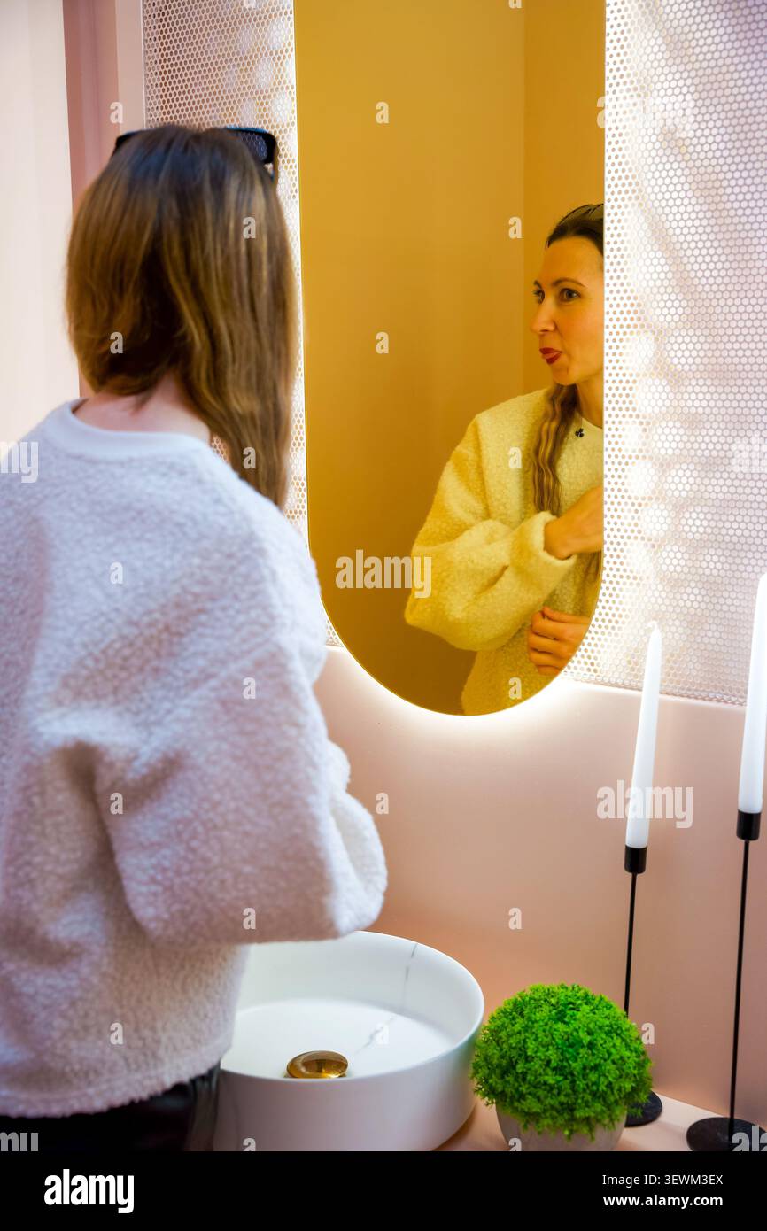 Femme faisant un visage drôle dans le miroir d'une salle de bain rose moderne avec un éclairage chaud Banque D'Images