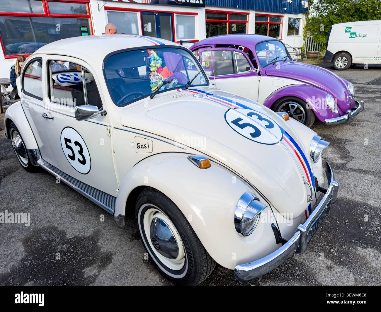 Un coléoptère volkswagen blanc décoré comme Herbie et un coléoptère volkswagen violet garé à l'extérieur d'un restaurant - Image de stock capturée avec un smartphone