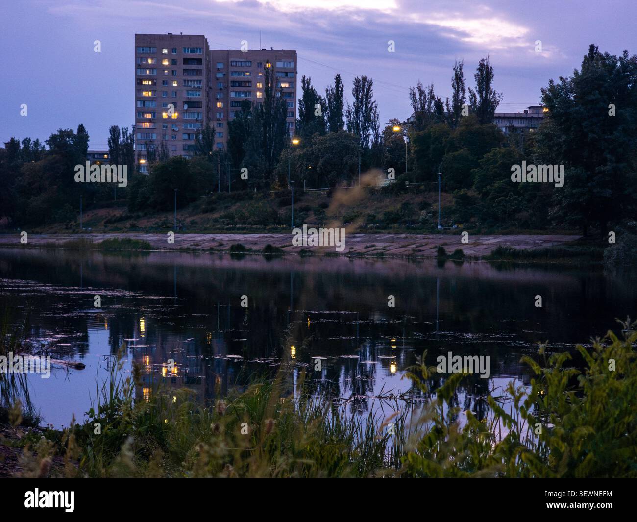 Bâtiments résidentiels bordant le canal de la rivière, leurs fenêtres éclairées projettent des reflets sur l'eau, avec des arbres et des lampadaires sur la rive du Banque D'Images