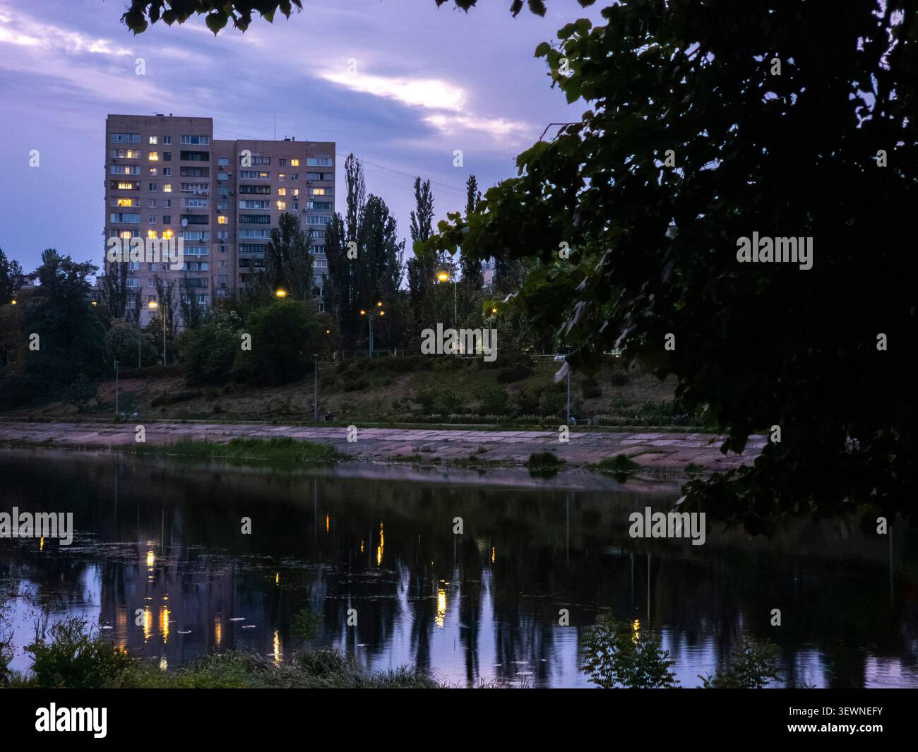 Bâtiment résidentiel de plusieurs étages se tenant au-dessus d'un canal de rivière calme avec des fenêtres éclairées reflétant sur la surface de l'eau après le coucher du soleil, encadré par d Banque D'Images