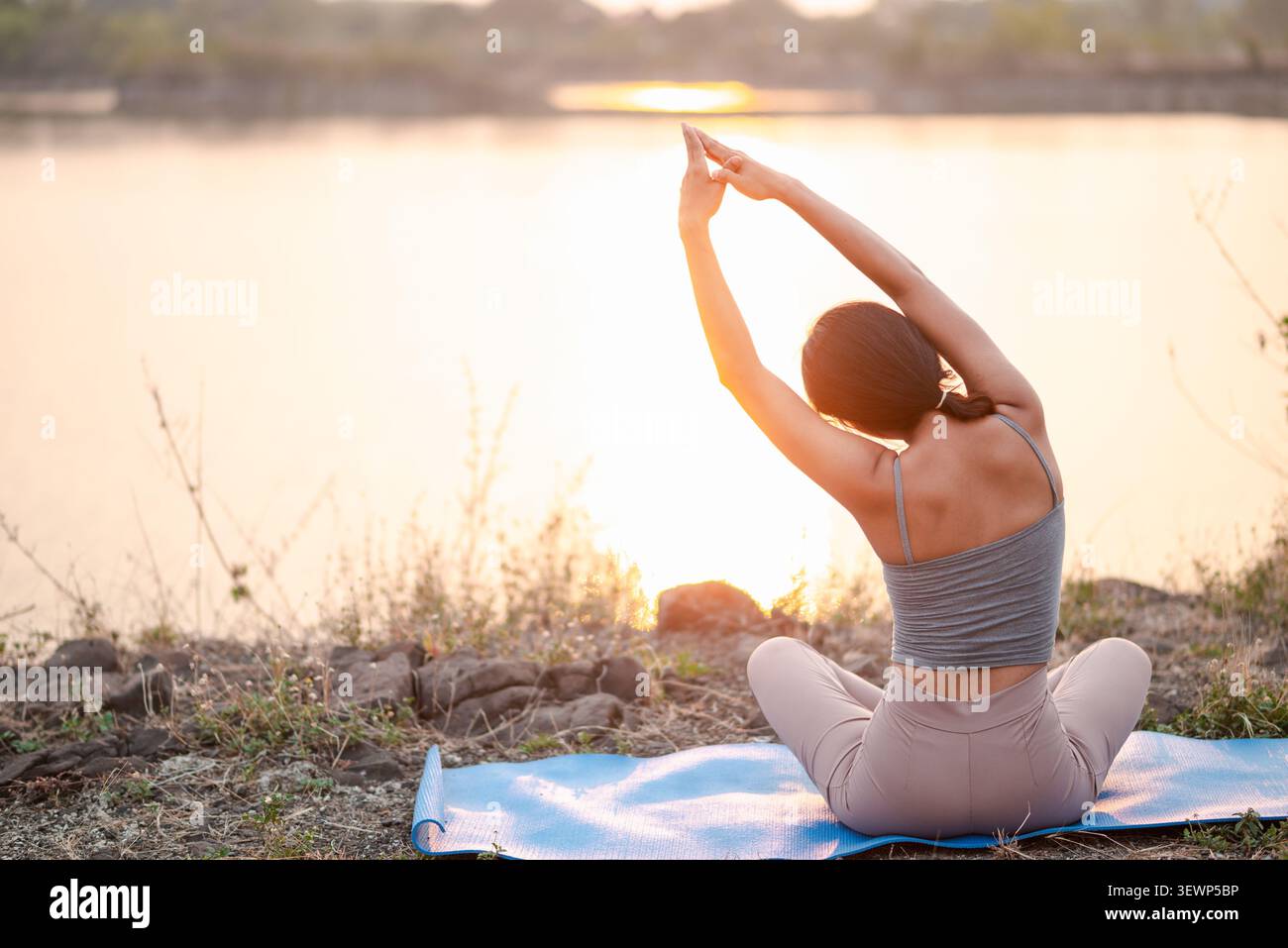 Vue arrière d'une femme asiatique assise sur un tapis de yoga bleu près d'un lac incandescent au coucher du soleil, bras étirés au-dessus de la tête, bien-être paisible, respiration consciente, outdo Banque D'Images