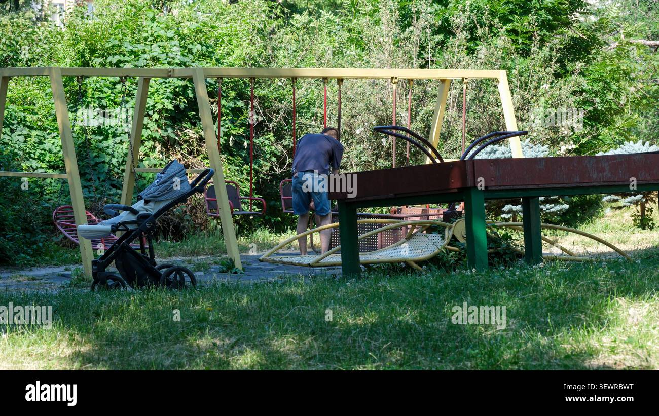 KIEV, UKRAINE - 28 JUIN 2026 : homme regardant son smartphone tout en se tenant debout près d'un enfant sur une aire de jeux dans le jardin botanique national M. M. Gryshko Banque D'Images