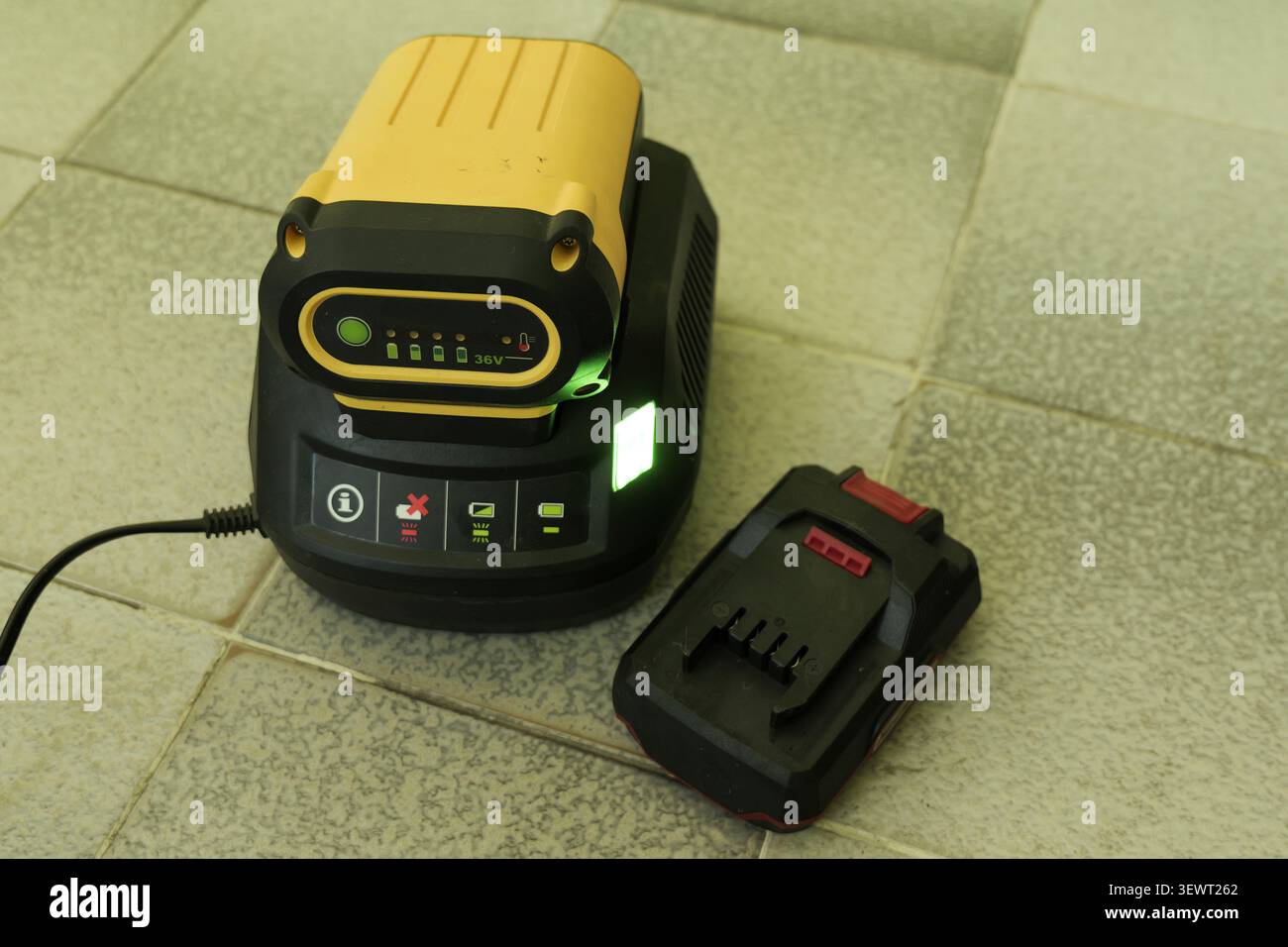 Batterie au lithium-ion jaune et noire se chargeant dans un chargeur d'outils électriques noir avec voyants lumineux. Un autre bloc-batterie reposant sur le plancher carrelé bes Banque D'Images