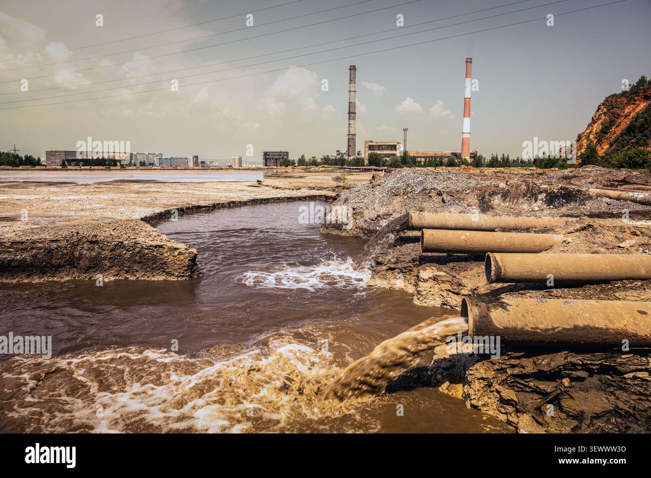 Les eaux usées sales s'écoulent des tuyaux de drainage dans un étang pollué près d'une zone industrielle. Une ville couverte de brume et de cheminées d'usine dans le backgro Banque D'Images