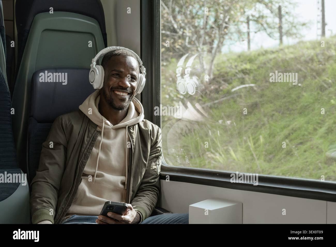 Un Afro-américain voyage en train, souriant et utilisant son smartphone et ses écouteurs tout en écoutant de la musique pendant son voyage. Banque D'Images