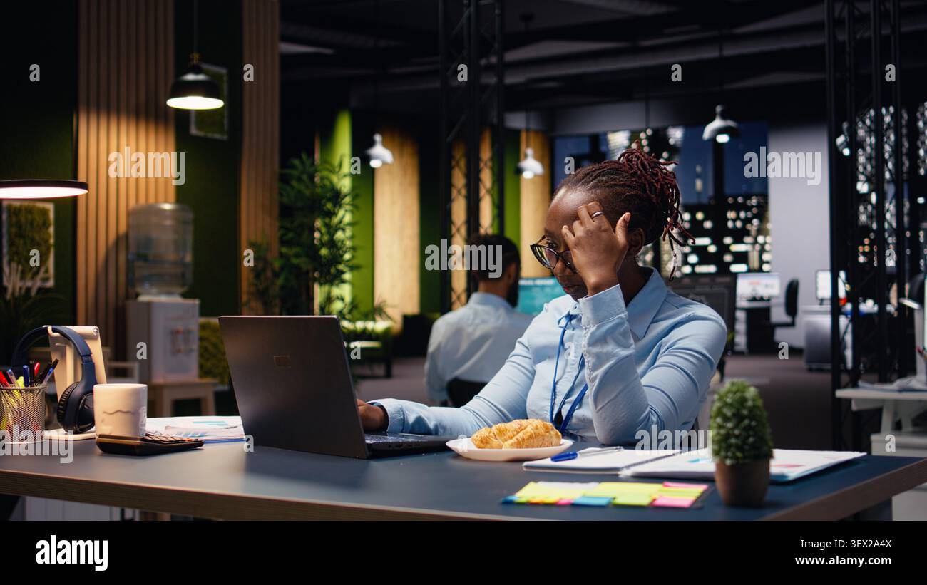 Trouble alimentaire et stress émotionnel, femme noire travaillant sur ordinateur portable, travailleuse mangeant trop de malbouffe à son bureau. Employé semblant inquiet alors qu'elle gère des tâches, surmanger compulsif. Banque D'Images