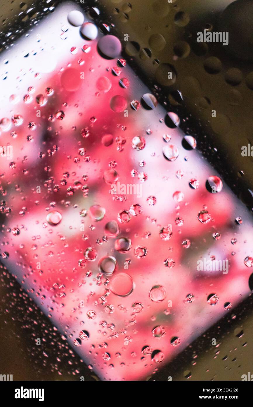 Des gouttes de pluie s'accrochent à une fenêtre, reflétant une lueur rouge floue. Les gouttelettes varient en taille et s'accrochent à la surface du verre. Lumière rose floue derrière la pluie su Banque D'Images