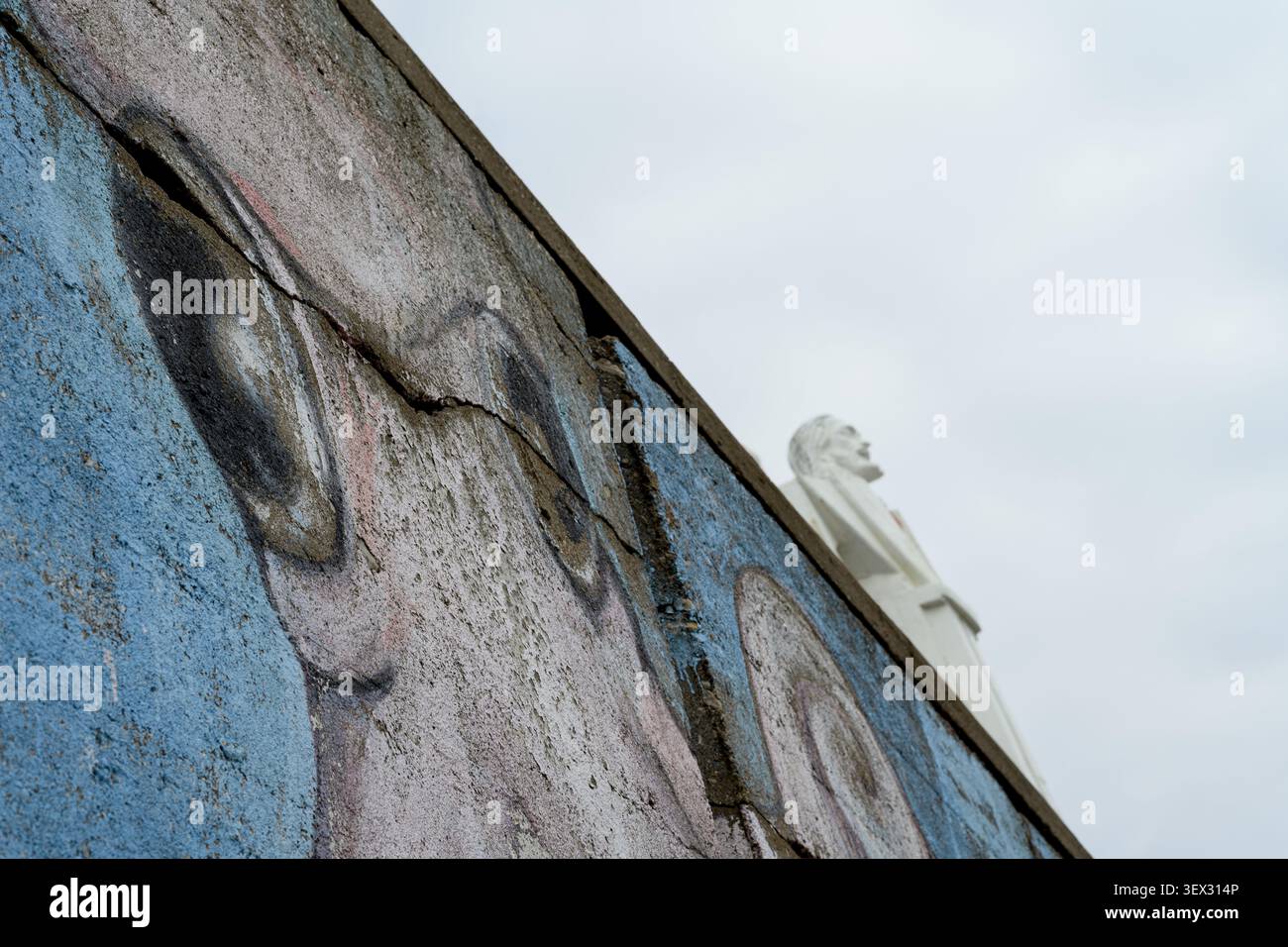 La murale altérée montre des graffitis décolorés sur un mur incliné. Le ciel gris apparaît au-dessus de la surface fissurée. Statue blanche se tient éloignée, silencieuse et austère Banque D'Images