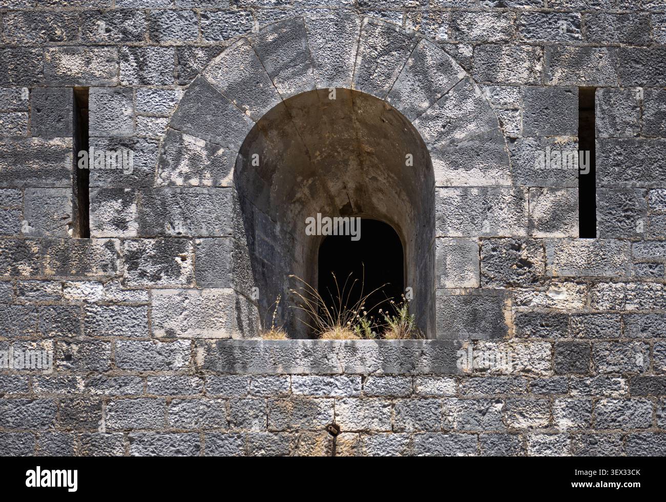 ancien mur de la forteresse en pierre avec une fenêtre d'embrasure voûtée et des fentes verticales pour flèches, avec des briques grises altérées et de l'herbe sèche poussant sur la led Banque D'Images