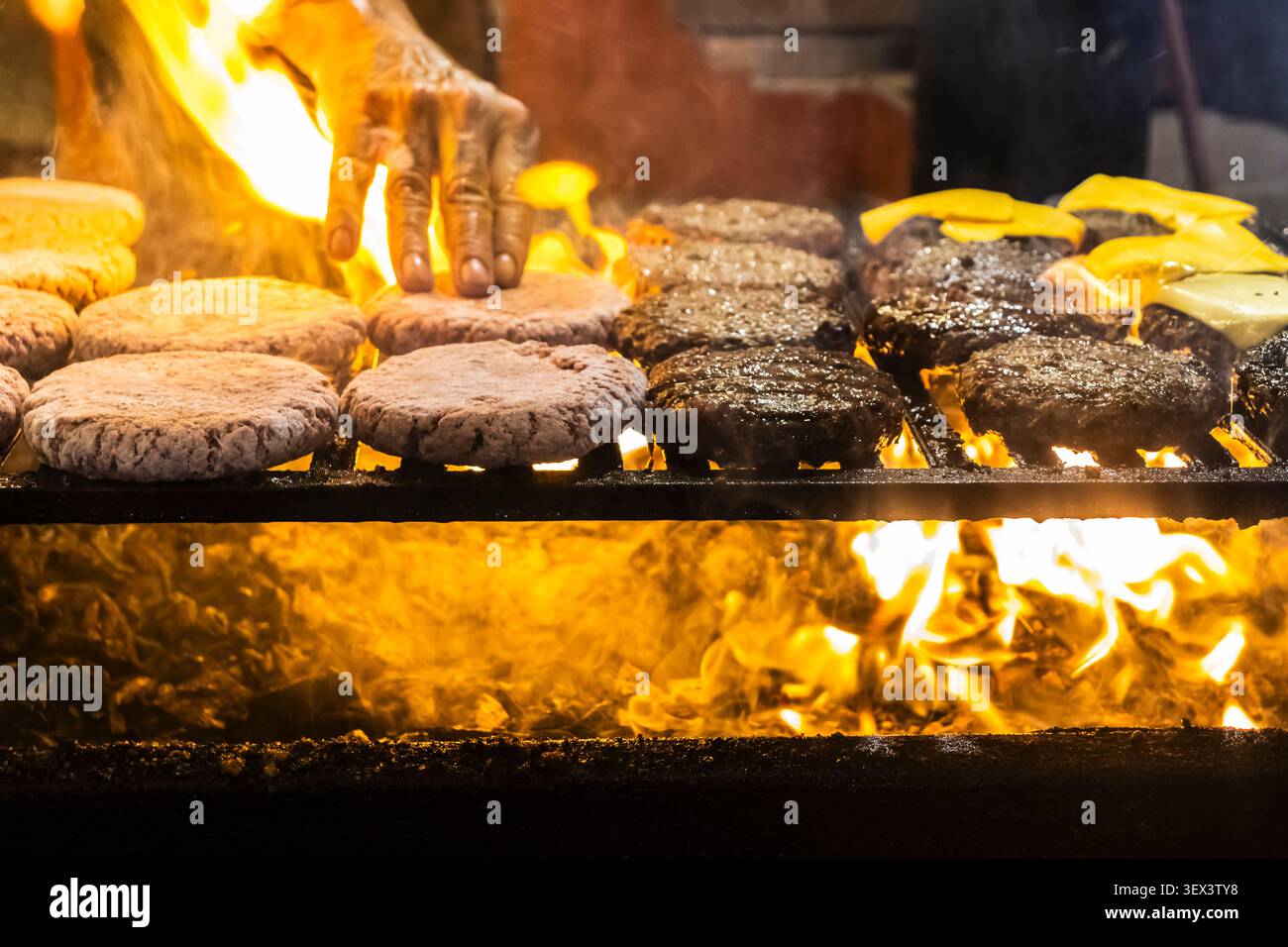 Gros plan sur les mains d'un chef disposant des galettes de viande crues et cuites sur une grille chaude avec des flammes rugissantes en dessous, préparant un hamburg artisanal traditionnel Banque D'Images