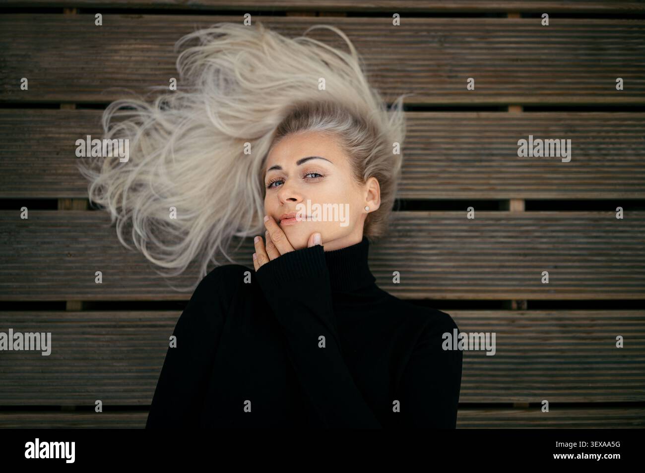 Belle femme blonde allongée sur un plancher en bois avec ses cheveux étendus, portant un col roulé noir Banque D'Images