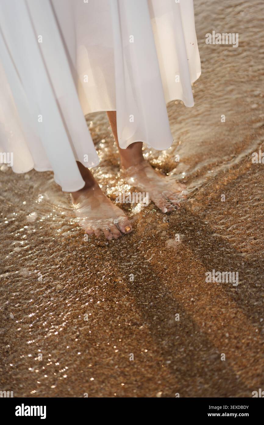 Femme pieds nus en robe blanche debout sur l'eau ondulations sur la plage de sable le matin Banque D'Images