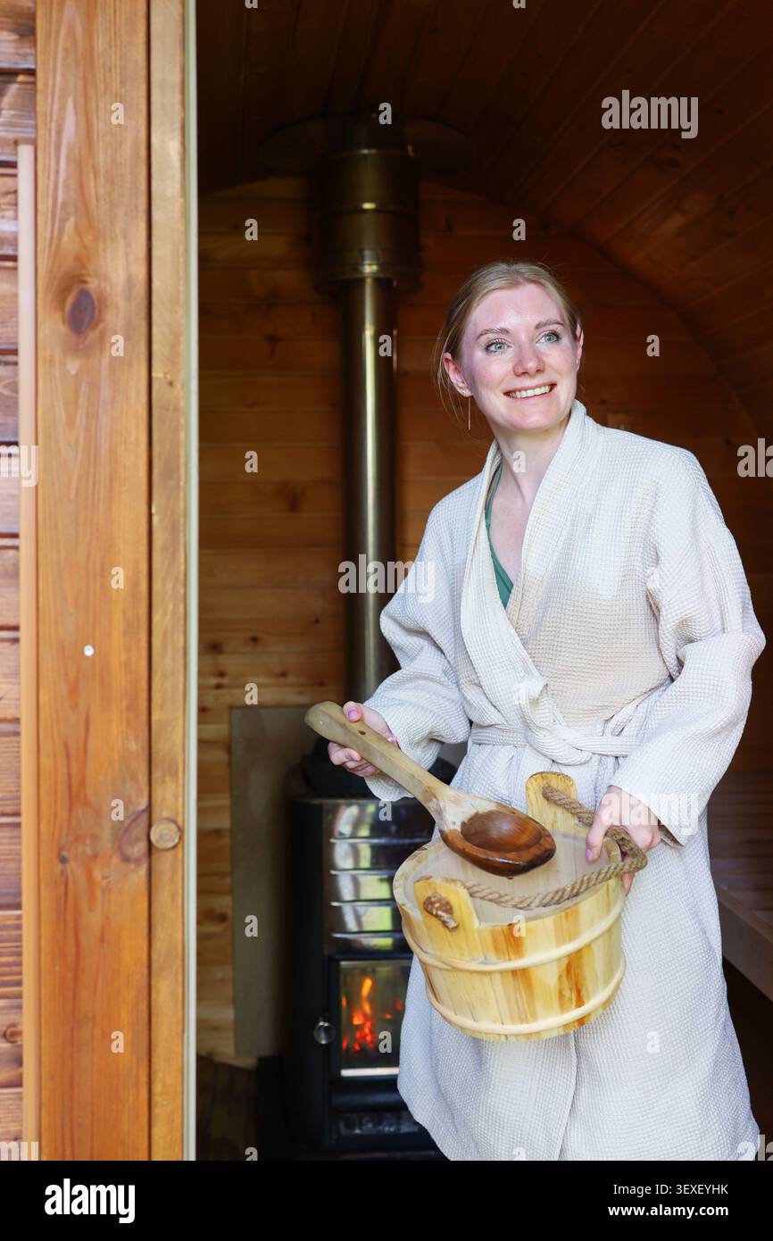 Femme souriante dans un peignoir tenant un seau en bois dans un sauna traditionnel, profitant d'une expérience de bien-être relaxante. Banque D'Images