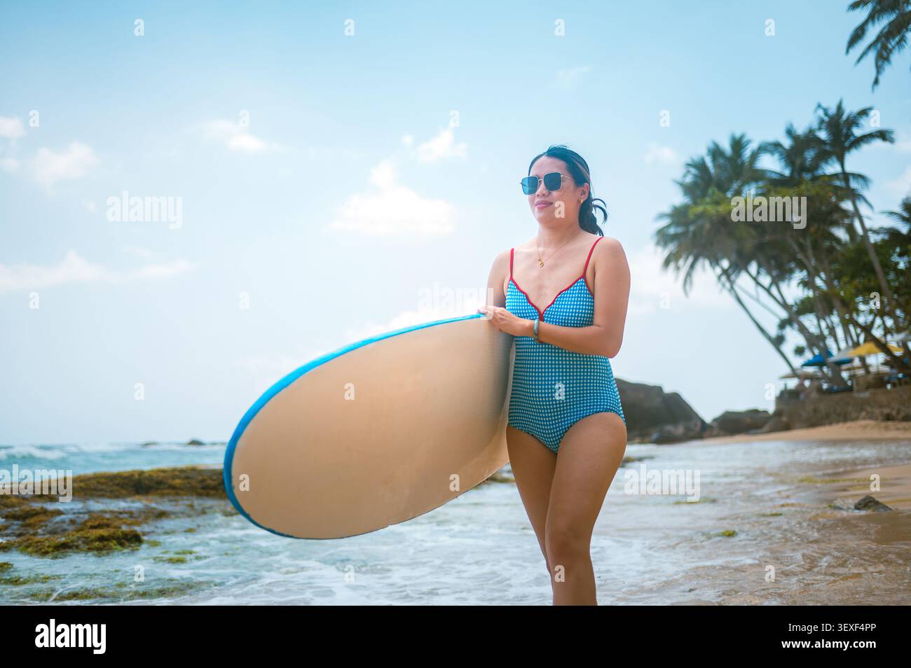 Une femme asiatique dans les années 30 se promène le long d'une plage pittoresque du Sri Lanka, tenant une planche de surf. Elle porte des lunettes de soleil et un maillot de bain élégant, avec un palmier Banque D'Images