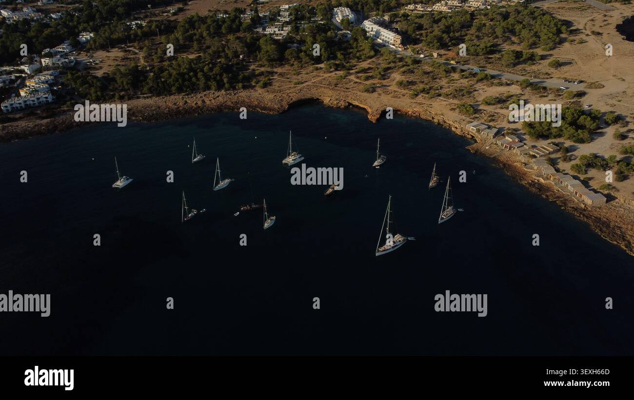 Vue drone de plusieurs voiliers ancrés dans une baie calme d'Ibiza avec des collines verdoyantes et des villages en arrière-plan Banque D'Images