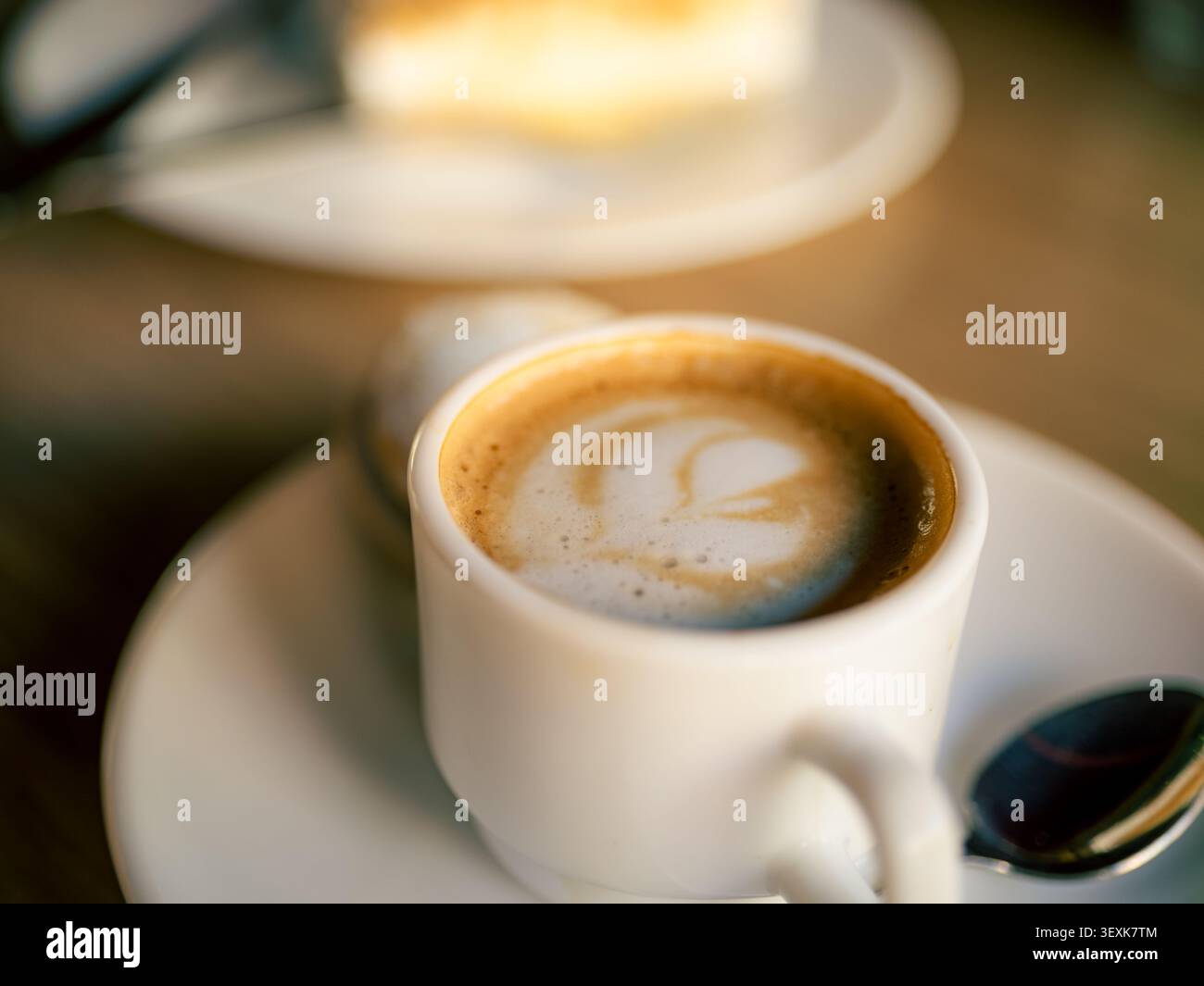 La tasse blanche peut contenir un latte crémeux Le lait mousseux tourbillonne dans le café. Cuillère repose sur une soucoupe à côté. Un arrière-plan doux montre une pâte floue. Cet im Banque D'Images