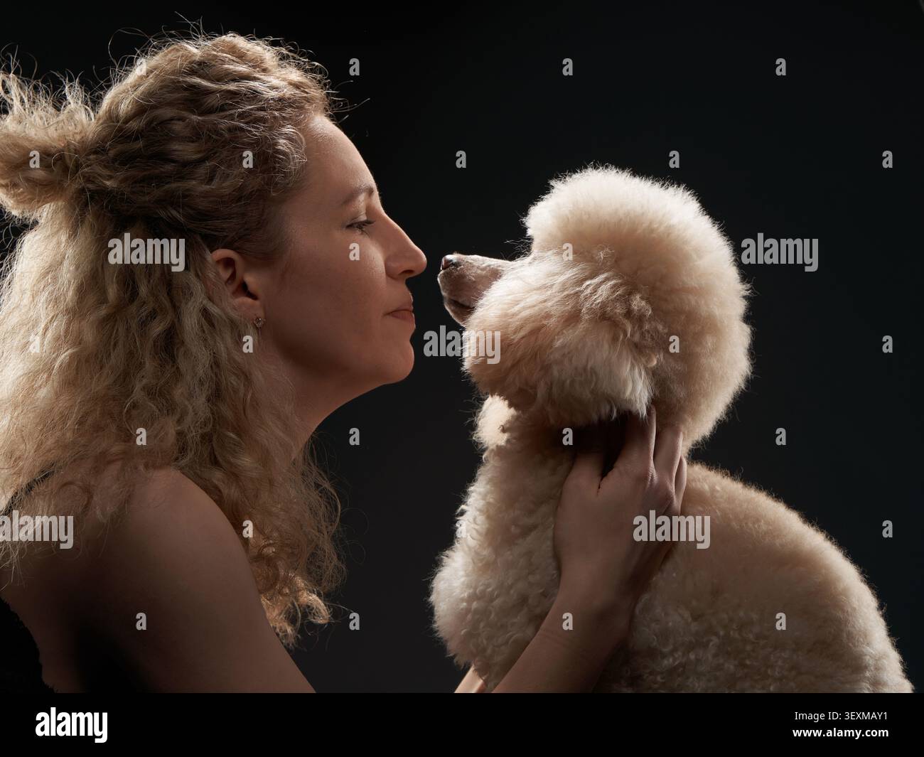 Une jeune femme blonde soulève avec amour un caniche blanc près de son visage. Le fond de studio noir intime améliore la connexion émotionnelle entre Banque D'Images