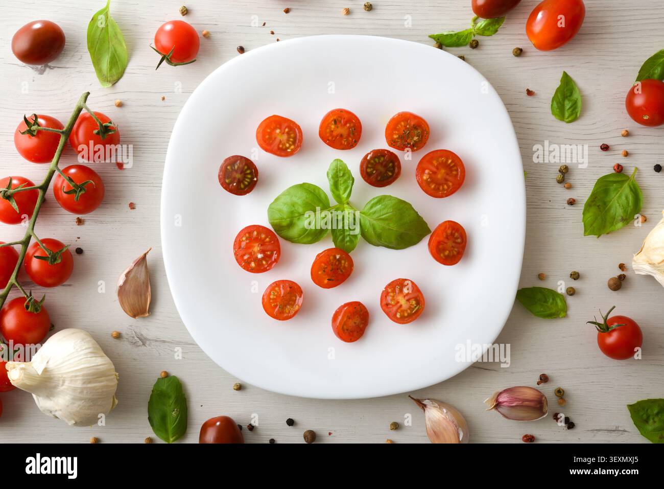Vue de dessus de tomates cerises tranchées avec basilic frais sur une assiette blanche, entourée d'ail, d'herbes et d'épices sur une table en bois rustique. Banque D'Images