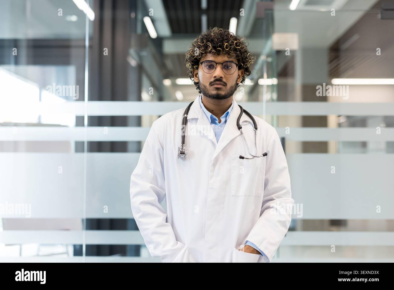 Jeune médecin indien avec des cheveux bouclés et des lunettes debout avec confiance dans un hôpital ou une clinique moderne, représentant des professionnels de la santé et la médecine Banque D'Images