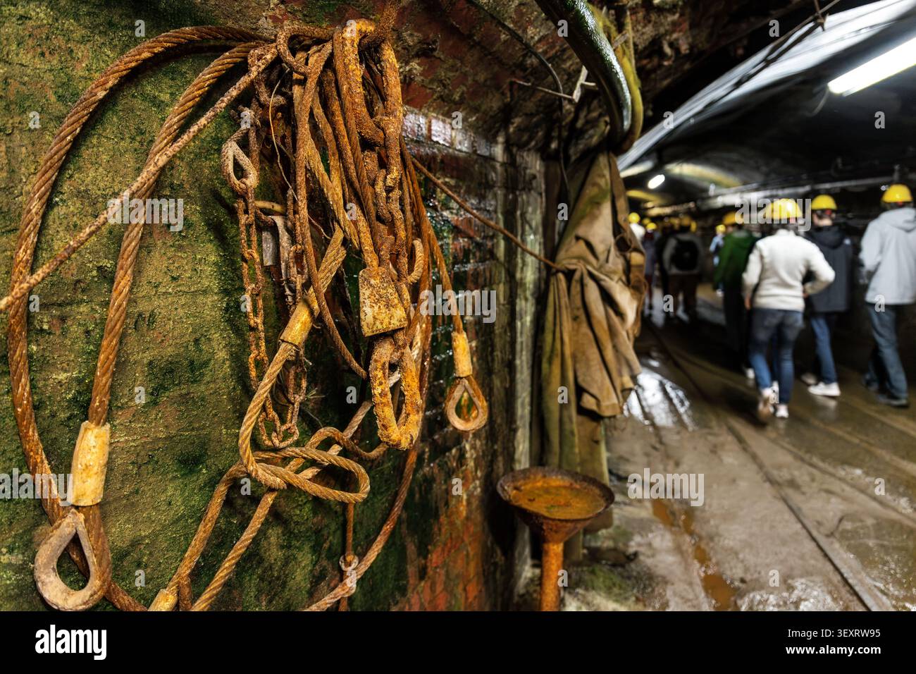 Des câbles et des crochets rouillés pendent sur un mur de briques humides à l'intérieur du tunnel de la mine tandis que les visiteurs casqués marchent le long du passage ferroviaire humide. Scène évoque le patrimoine industriel Banque D'Images