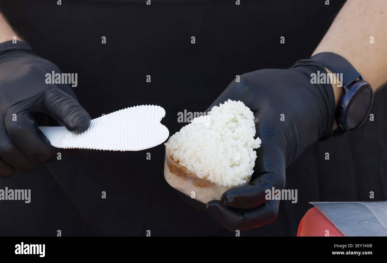 Mains d'un cuisinier portant des gants d'hygiène noirs pressant du riz blanc collant dans un moule triangulaire pour faire des onigiri. Préparation de la cuisine de rue asiatique au F Banque D'Images