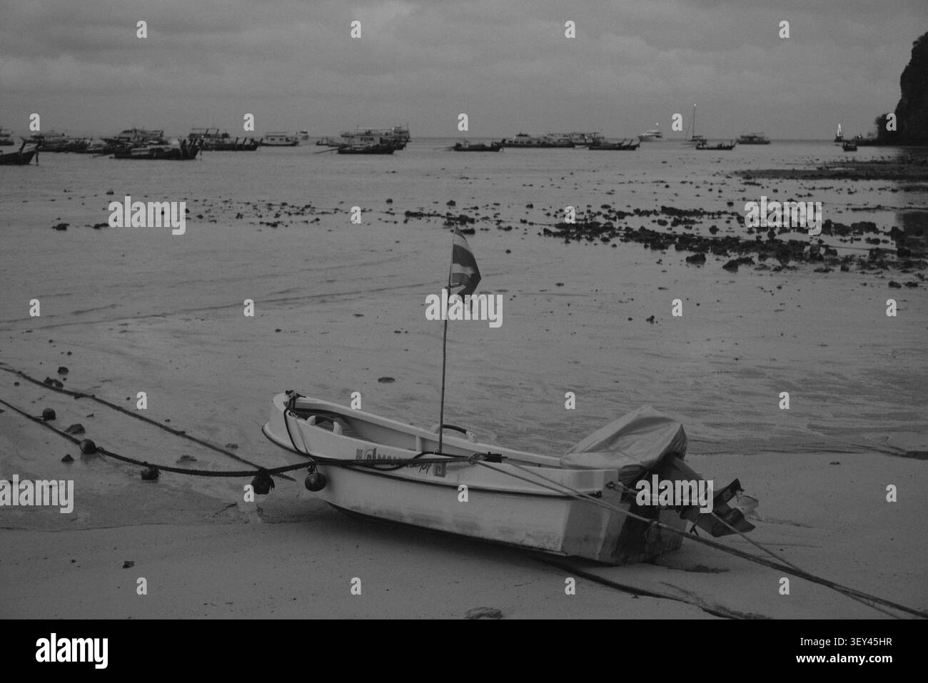 Îles Phi Phi, Thaïlande – 18 mai 2026 : un petit bateau à moteur repose sur le rivage sablonneux exposé à marée basse, avec des bateaux thaïlandais traditionnels à longue queue à l’ancre Banque D'Images