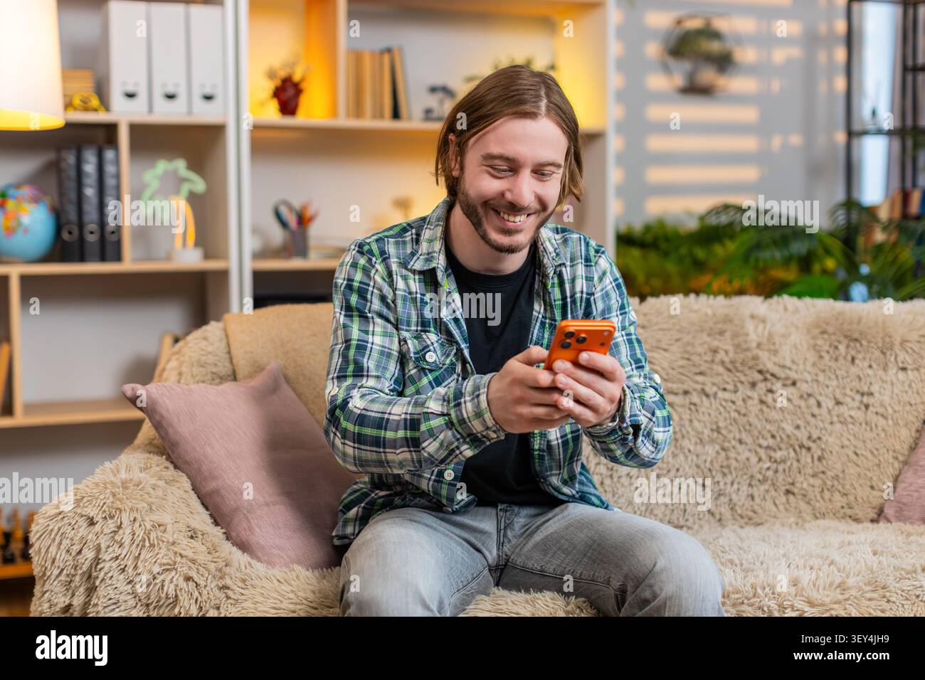 Jeune homme sur le canapé de la maison lit le message drôle de smartphone sourit avec une surprise chaude et commence à rire. Mec adulte sur le canapé de la maison aime le cha mobile joyeux Banque D'Images