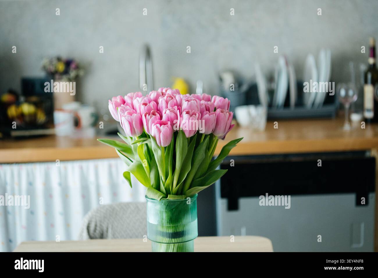Un bel arrangement de tulipes roses dans un élégant vase vert, placé sur une table à manger en bois dans un intérieur confortable de cuisine à domicile. Banque D'Images