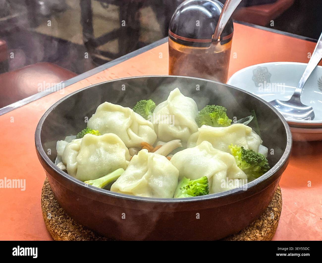Un bol en pierre noire rempli de boulettes chinoises bouillantes fumantes et de fleurettes de brocoli, photographié dans un cadre de restaurant avec de la vapeur visible s'élevant du plat chaud. Banque D'Images