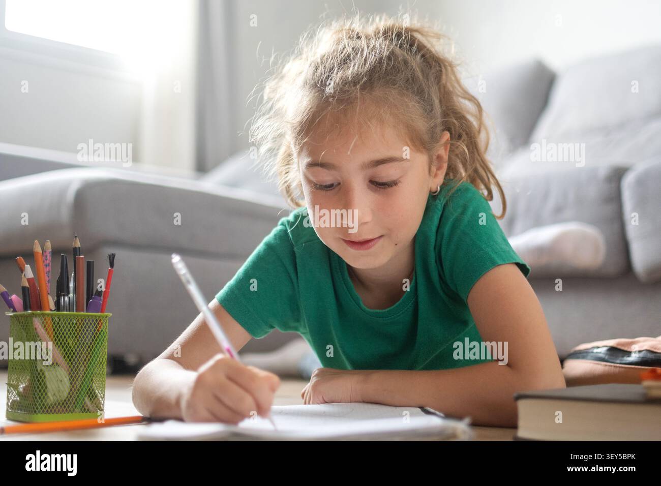 Fille de l'école primaire écrivant des devoirs tout en étant allongée sur le plancher du salon à la maison Banque D'Images