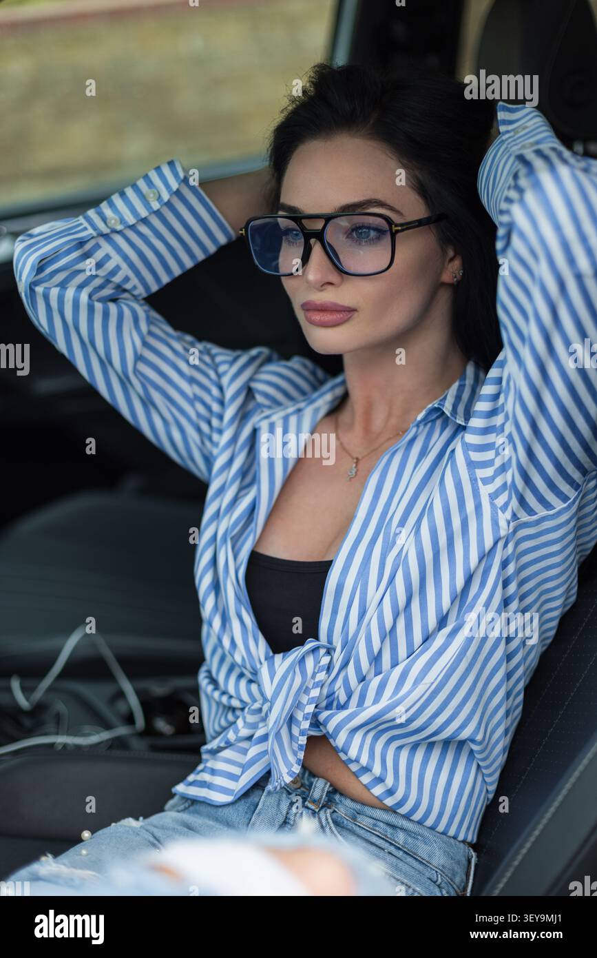 Une belle femme brune portant des lunettes d'aviateur à la mode se détend dans un siège d'auto, passant ses mains dans ses cheveux et regardant loin réfléchi. Cas Banque D'Images