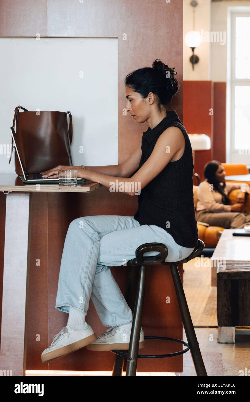 Vue latérale de la femme d'affaires concentrée travaillant sur son ordinateur portable tout en étant assis sur le tabouret à l'espace de coworking Banque D'Images