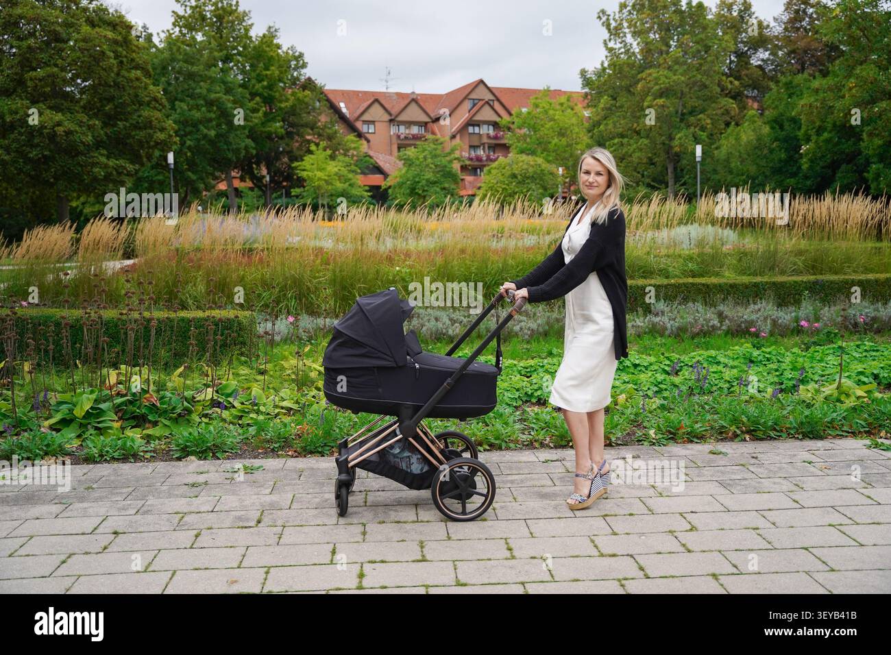 Maman d'affaires vêtue d'une robe blanche sur sa pause déjeuner poussant une poussette de bébé noir sur un jour d'automne lumineux dans le parc, maman est une blonde européenne Banque D'Images