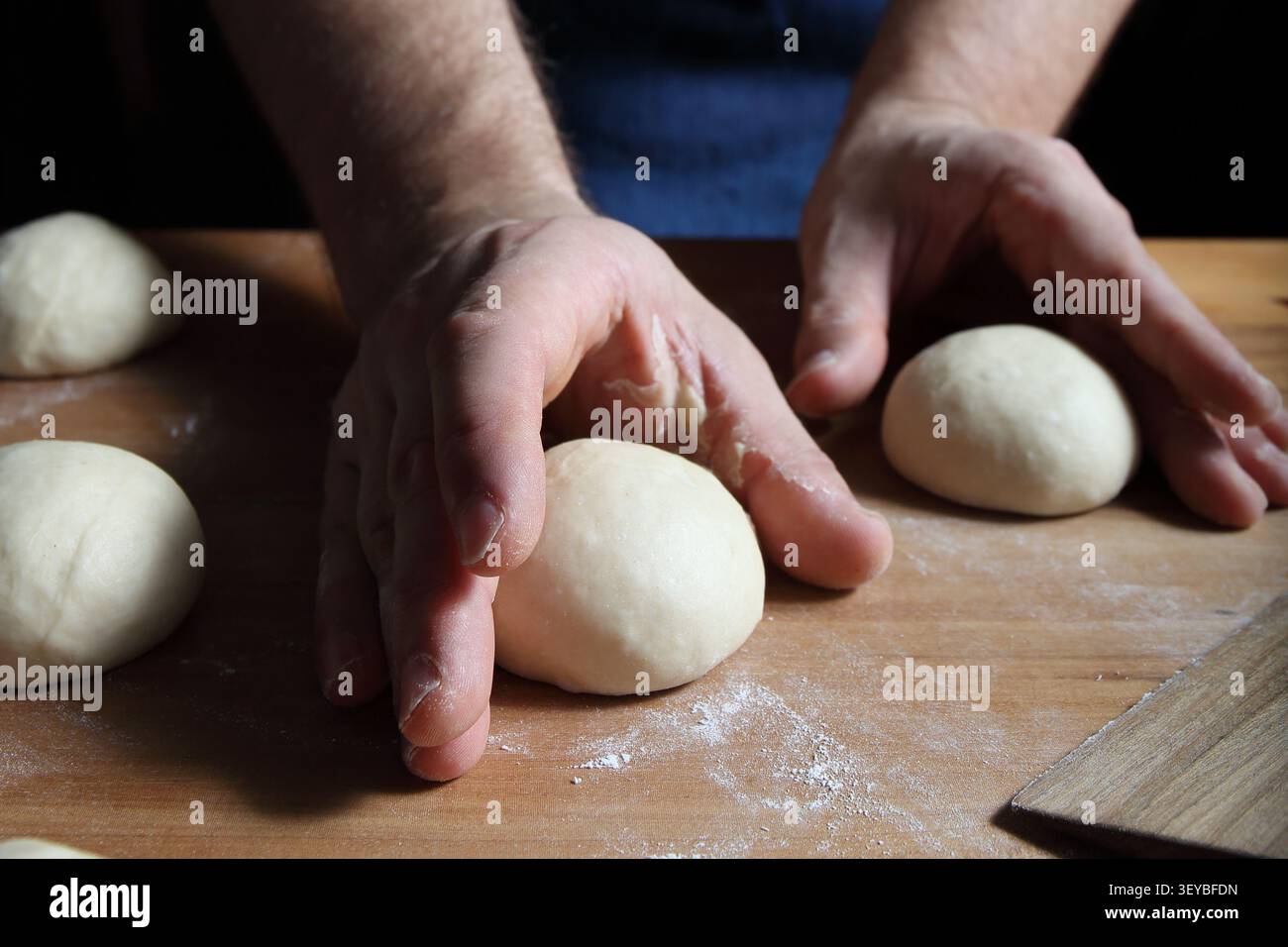 Mains façonnant des boules de pâte lisses sur une planche de bois farinée avant l'épreuve. Préparation de petits pains hamburger faits maison ou de petits pains dans une cuisine à domicile. Banque D'Images